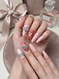 A close-up of elegant hands wearing luxury pink and red press-on nails with Dior monogram pattern, crystal charms, and bows, with a Miss Dior perfume bottle in the background.